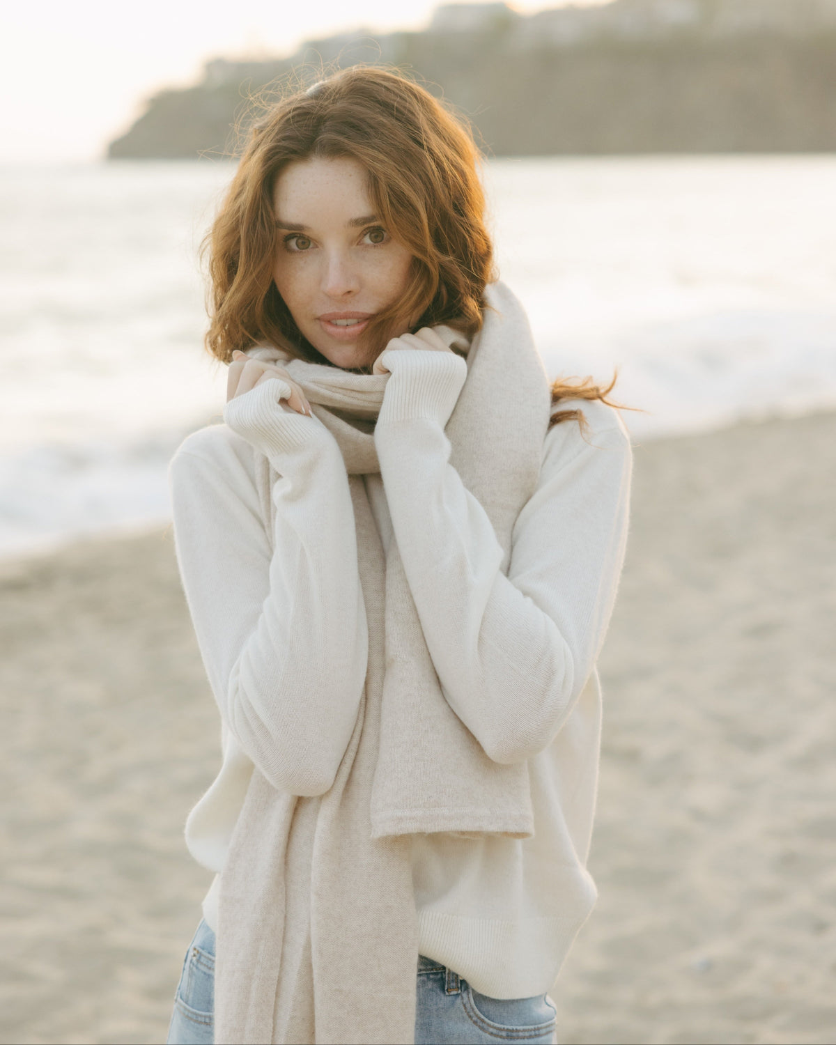 Woman wearing a white sweater and 
ultra soft wool/cashmere blend wrap scarf   on a beach. Cashmere Travel Wrap in Oatmeal by Ellsworth + Ivey.