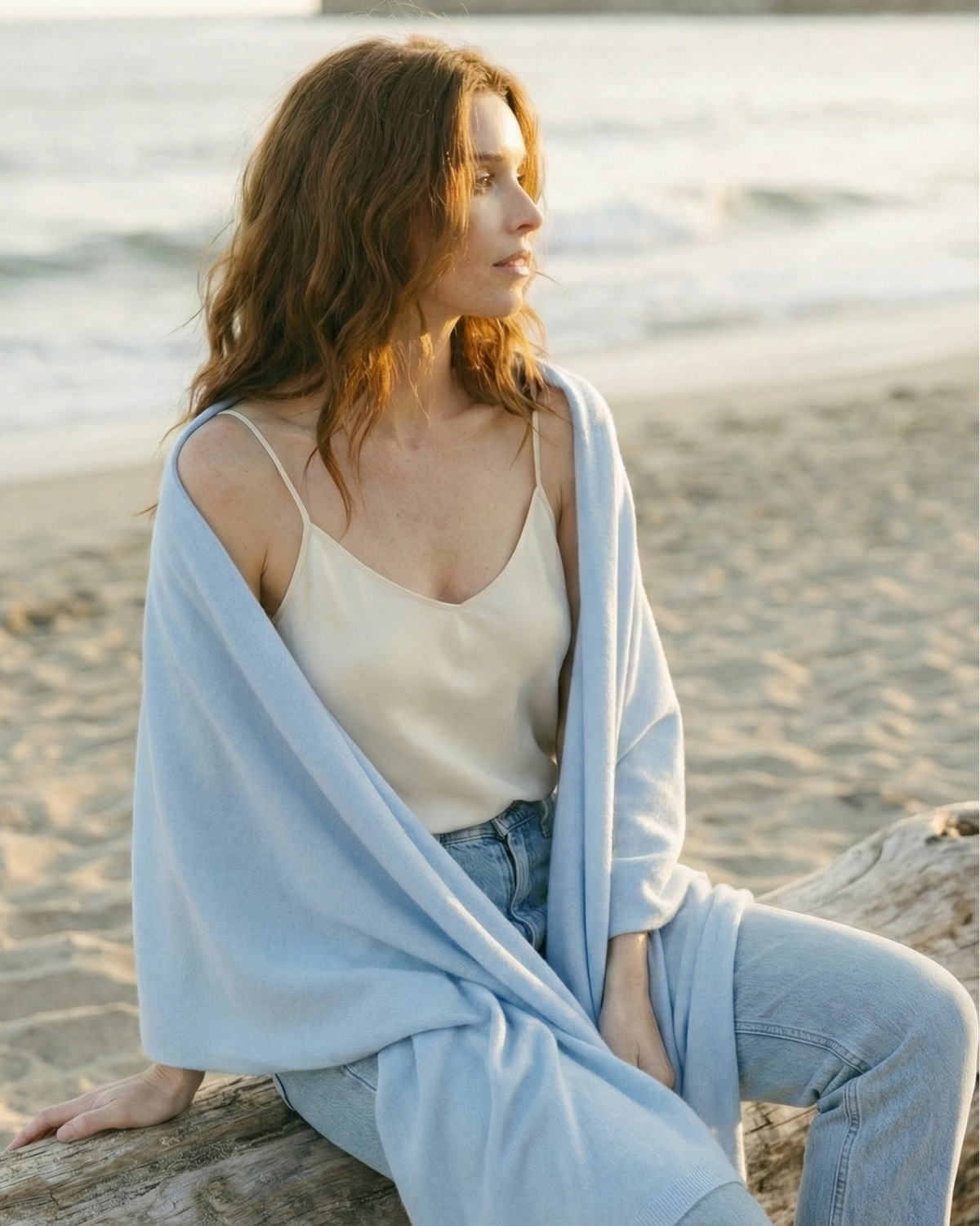 Woman wearing an ultra soft sky blue wool/cashmere blend wrap scarf on a beach. Cashmere Travel Wrap in Sky Blue by Ellsworth + Ivey.