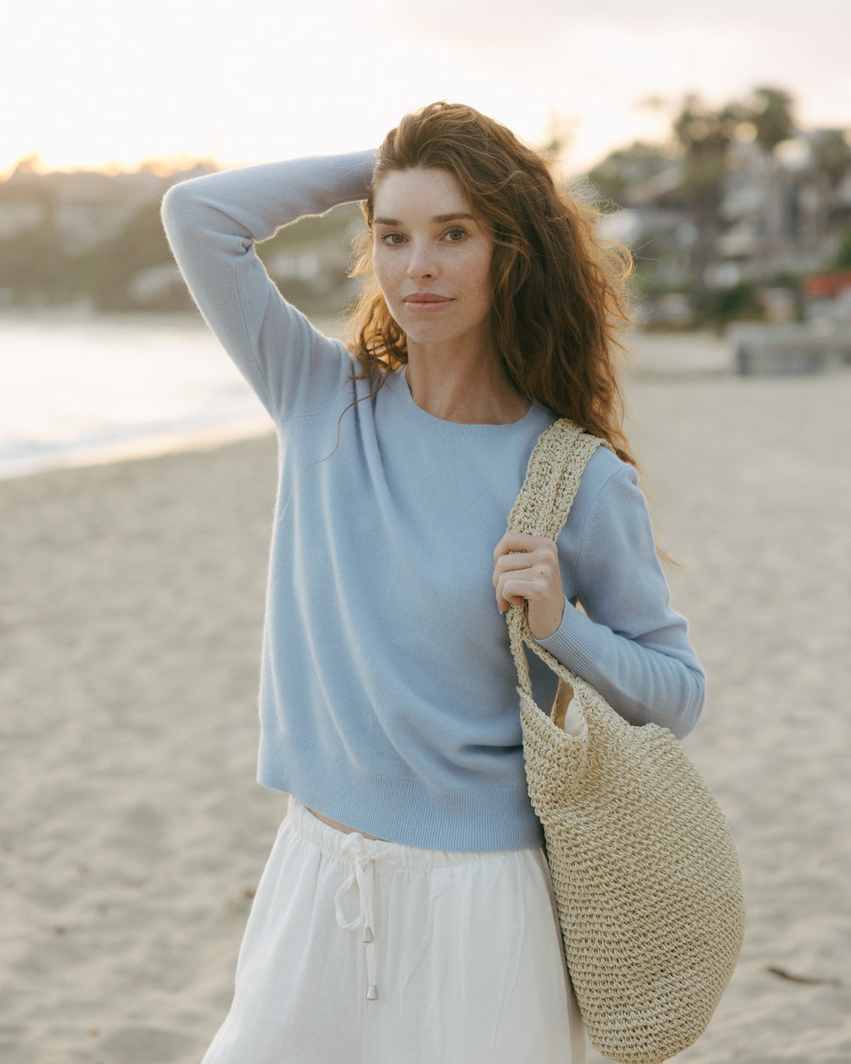 Woman on a beach holding a woven bag, wearing a light blue crewneck cashmere women's sweater and white pants. Daphne Crew Sweater in Sky Blue by Ellsworth + Ivey.