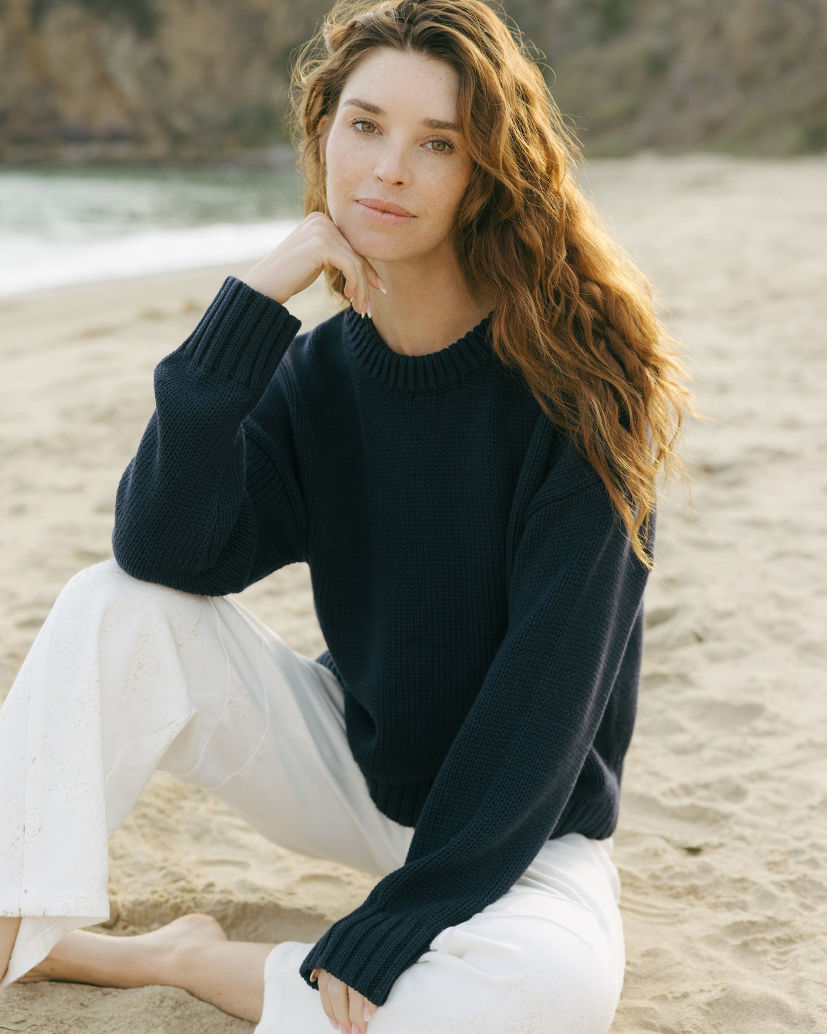 Woman sitting on a beach wearing a navy blue sweater and white pants. Elena Crew Sweater in Navy by Ellsworth + Ivey.