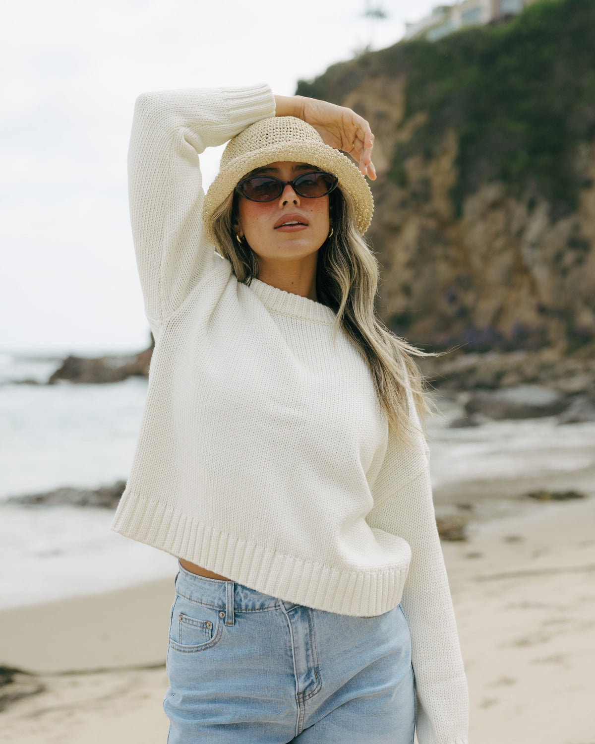 Woman on a beach wearing a white sweater, straw hat, and sunglasses with a blurred background. Elena Crew Sweater in Cornflower Ivory by Ellsworth + Ivey.