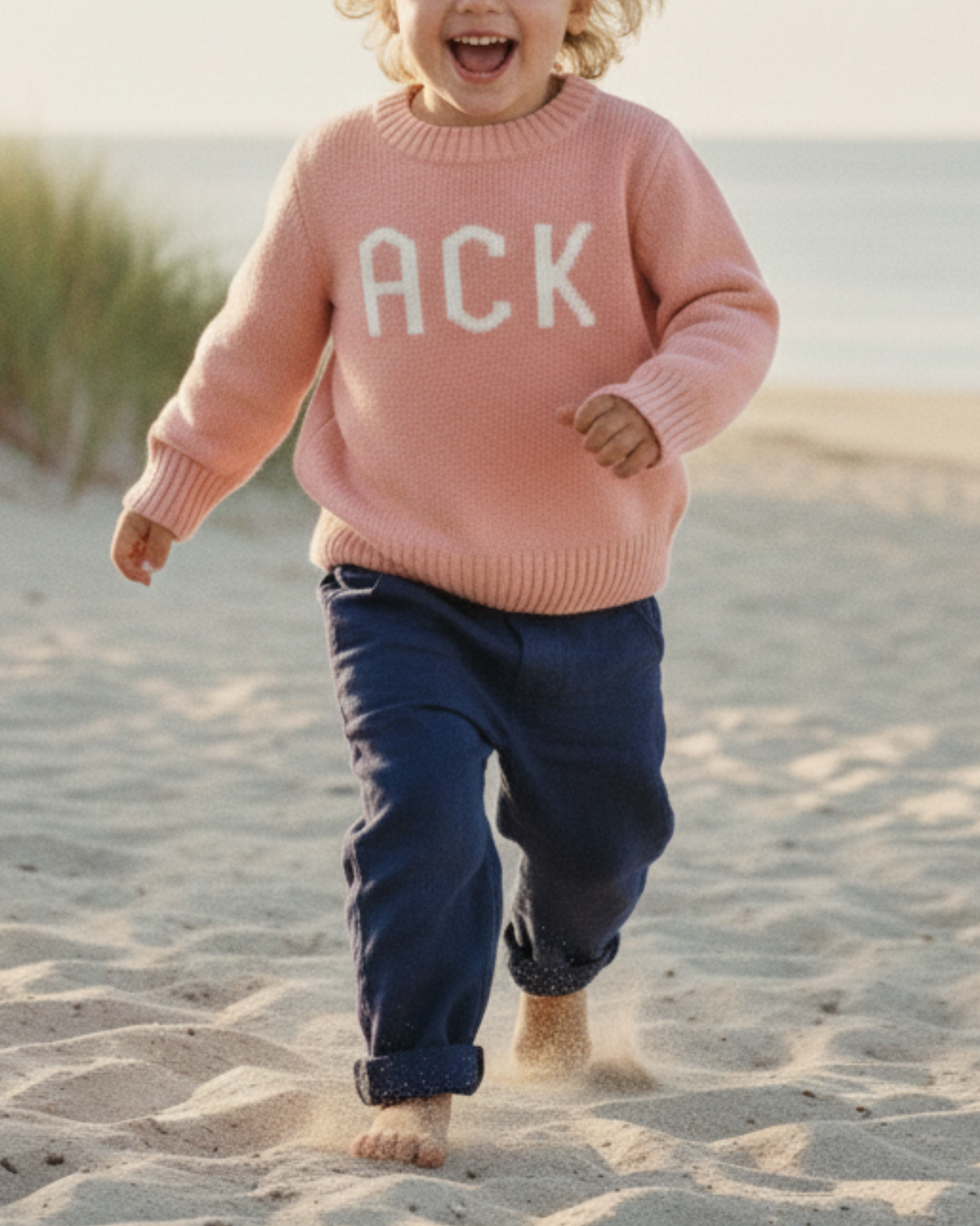 Child running on a sandy beach wearing a salmon sweater with 'ACK' on it.