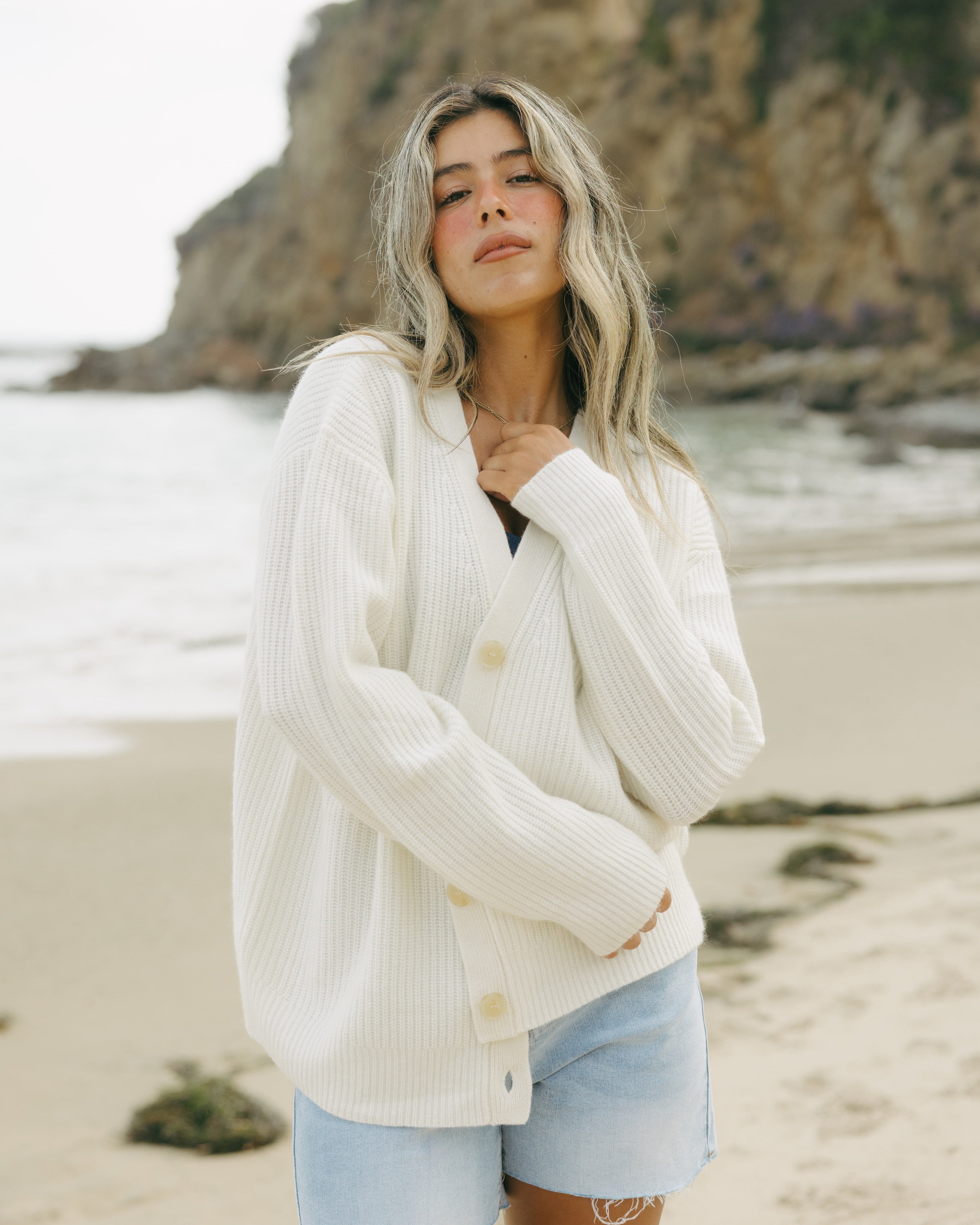Woman wearing an ivory off-white v-neck oversized cashmere women's cardigan and denim shorts on a beach with a scenic background. Lilah V-Neck Knit Cardigan in Ivory by Ellsworth + Ivey.