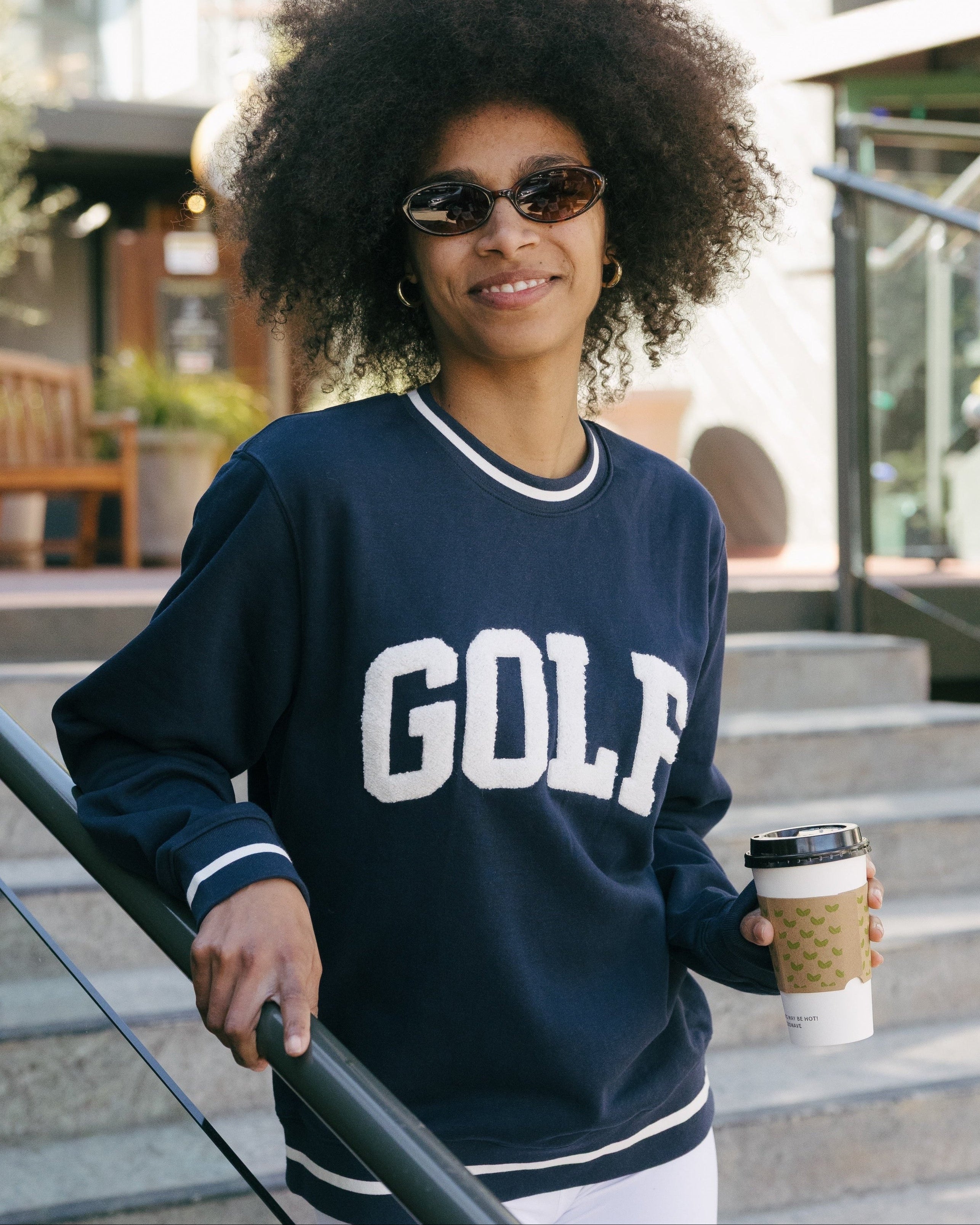Woman wearing a navy 'GOLF' sweatshirt holding a coffee cup outdoors. Elle Sweatshirt - Collegiate Golf by Ellsworth + Ivey.
