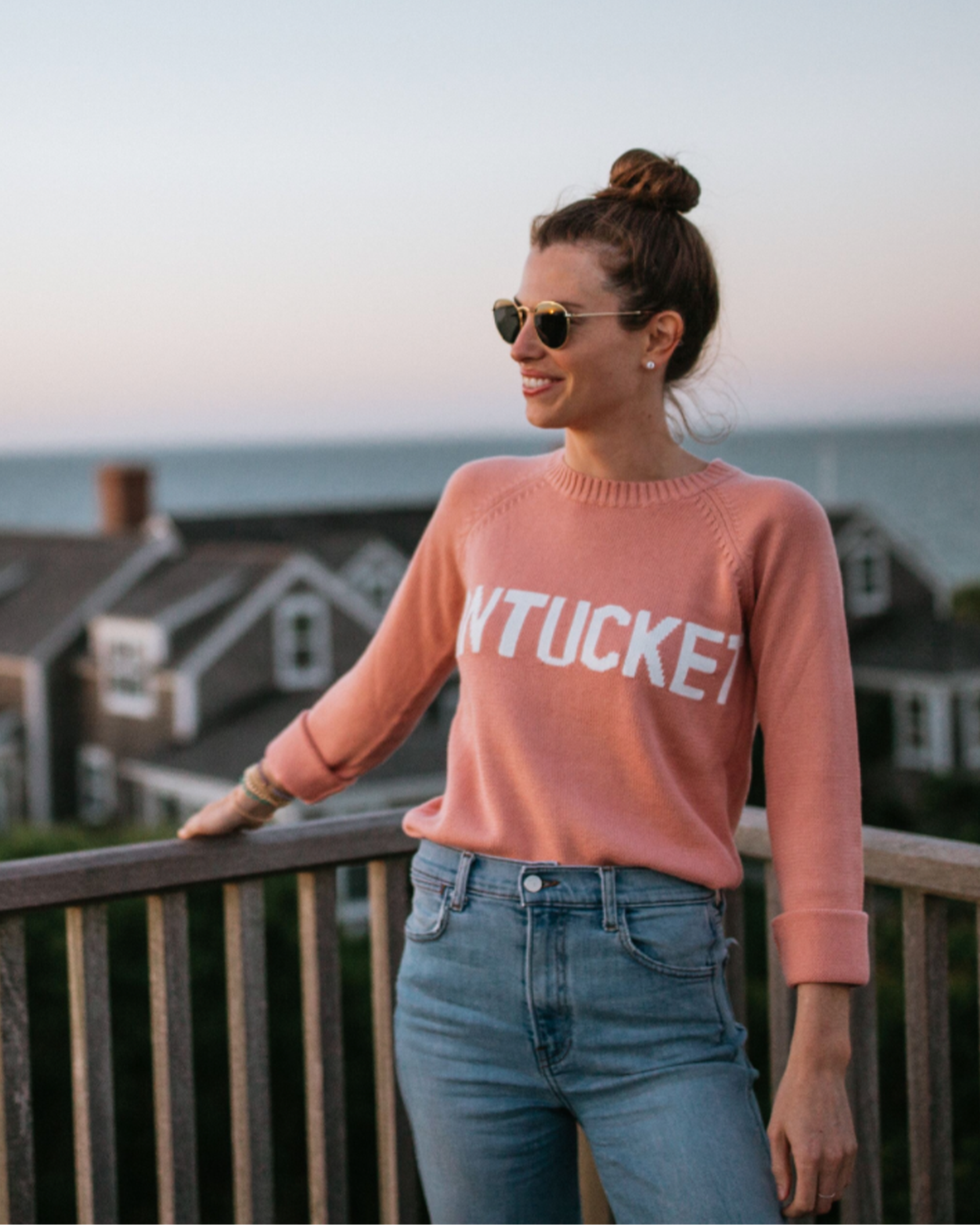 Women wearing a salmon peach pink sweater with white 'Nantucket' across the chest, outside. Classic Crew Sweater- Nantucket by Ellsworth + Ivey.